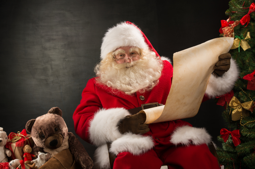 Portrait of happy Santa Claus sitting at his room at home near Christmas tree and big sack and reading Christmas letter or wish list